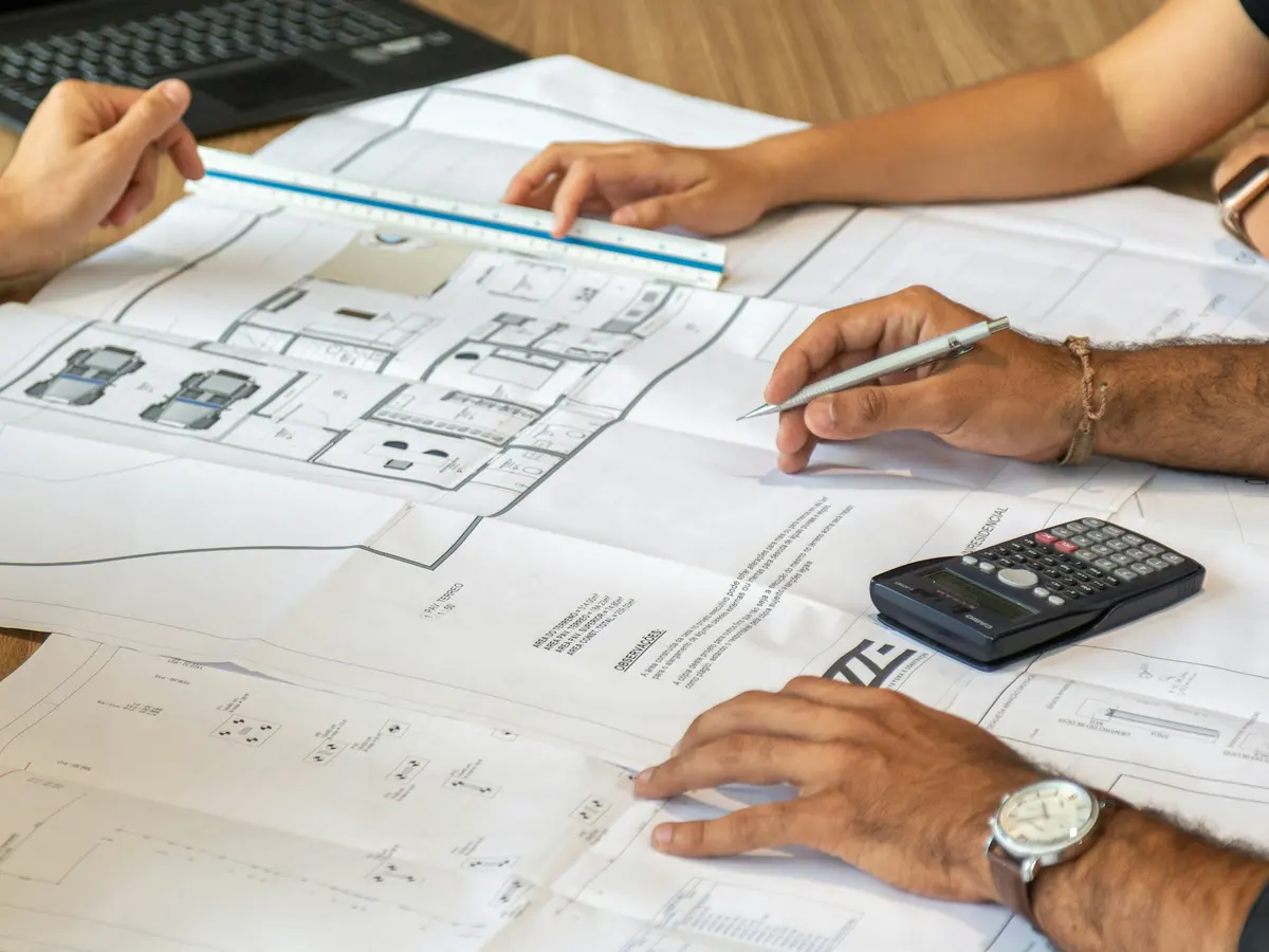 Mãos revisando pranchas de projeto com calculadora sobre a mesa